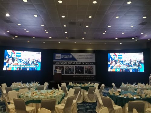 Salón con mesas redondas para banquete corporativo con dos pantallas LED laterales y back central impreso con el branding del Logistics World Summit.