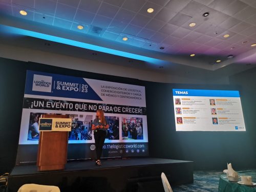 Oradora en podio dando una ponencia en el LOGISTICS WORLD SUMMIT, con dos pantallas LED de fondo mostrando el branding y la agenda.