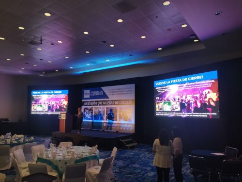Salón con mesas redondas para cena de gala, con triple pantalla LED mostrando promoción de evento social de cierre del Logistics World Summit.