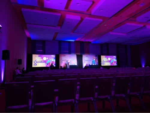 Salón de convenciones con montaje de escenario multipantalla e iluminación ambiental morada para congreso corporativo masivo.