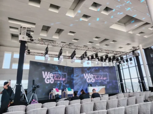 Salón de conferencias amplio durante un congreso corporativo masivo, con montaje tipo auditorio lleno de asistentes, grandes pantallas de proyección en el escenario e iluminación ambiental azul y morada.