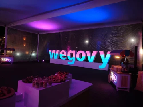Montaje de evento nocturno con letras gigantes iluminadas de la marca Wegovy, iluminación ambiental azul y morada bajo carpa y decoración tipo lounge.