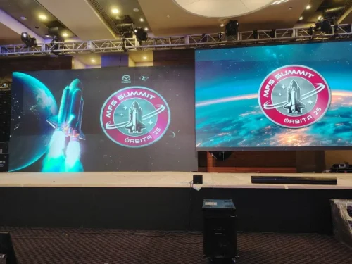 Escenario de evento corporativo con dos pantallas LED gigantes mostrando gráficos espaciales de un cohete y logotipos del evento.