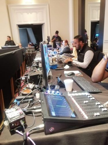 Mesa de control audiovisual con consola de mezcla y staff técnico operando computadoras y monitores en un evento corporativo.