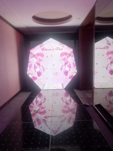 Pantalla LED con forma de hexágono iluminada en color rosa y blanco ubicada al final de un pasillo con piso negro reflectante.