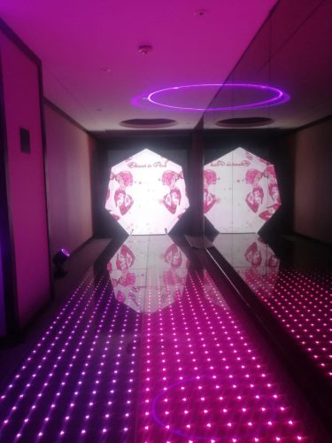 Túnel inmersivo iluminado en color rosa con piso de luces LED y espejos laterales en entrada de evento corporativo.