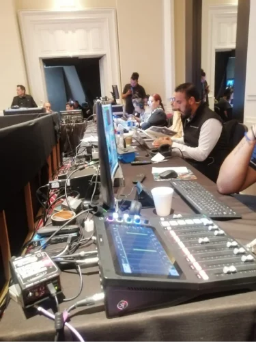Mesa de control técnico con consola de audio digital y staff operando computadoras durante un evento corporativo en vivo.