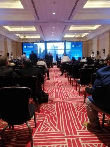 Salón de hotel con montaje de sillas tipo auditorio para conferencia corporativa, mostrando audiencia sentada y pantallas al fondo.